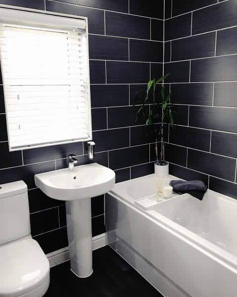 Modern bathroom with black tiled walls, white bathtub, pedestal sink, and green plant decor.