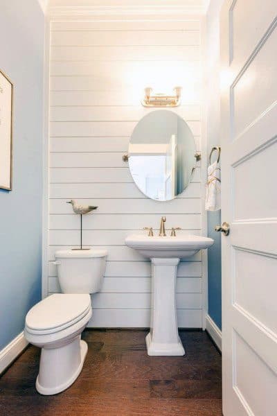 A cozy half bath with a round mirror, pedestal sink, toilet, wood floor, and light blue shiplap walls