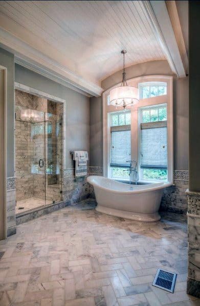 Elegant bathroom with freestanding white bathtub, marble herringbone floor, and glass shower.