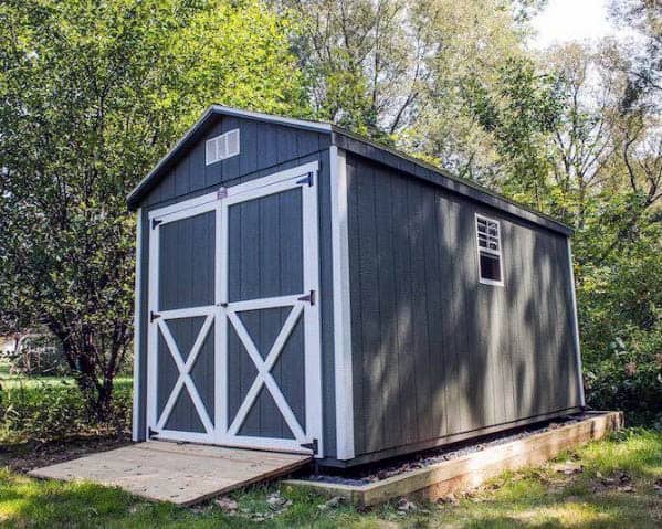 Dark gray barn-style shed with white double doors and a small ramp, set in a wooded backyard.