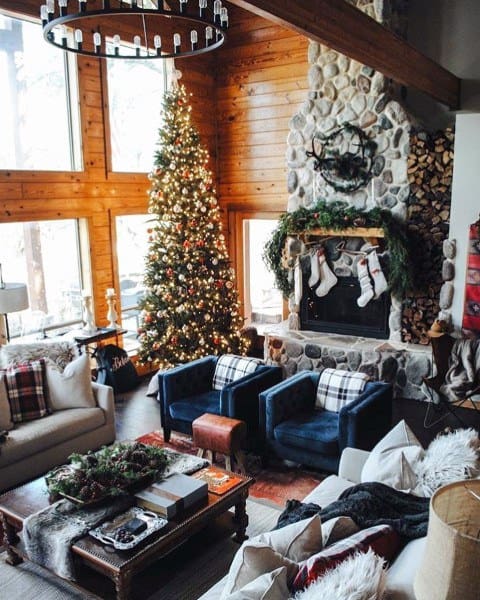 A cozy, traditional rustic living room featuring a stone fireplace, large windows, a Christmas tree, and festive decorations, creating a warm holiday atmosphere