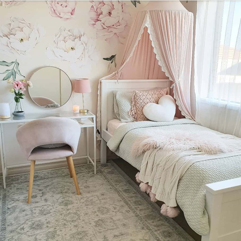 A cozy pastel pink bedroom with a canopy bed, floral wallpaper, and a small vanity table with a mirror and chair