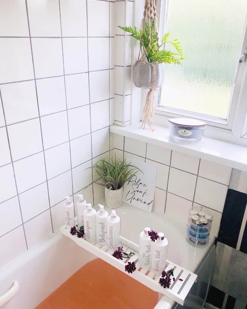 White tiled bathroom with a bathtub tray holding bottles and hanging plant near window.