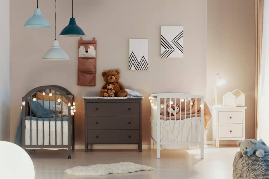 Baby room for twins with two cribs, string lights, modern wall art, and cozy lighting fixtures.