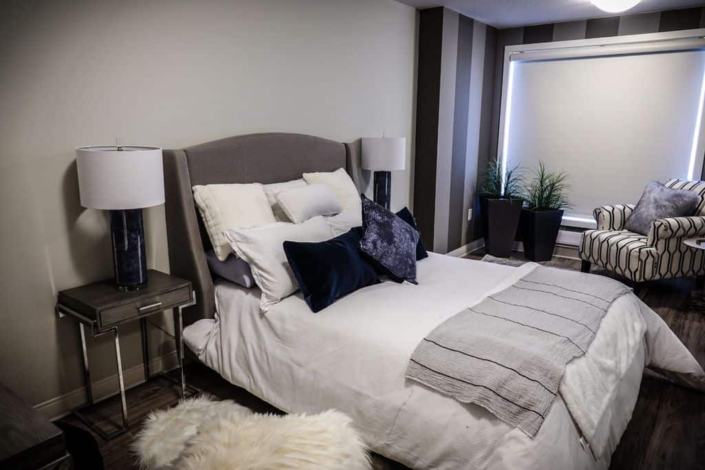 Elegant bedroom with striped walls, gray bed, potted plants, and a patterned lounge chair.