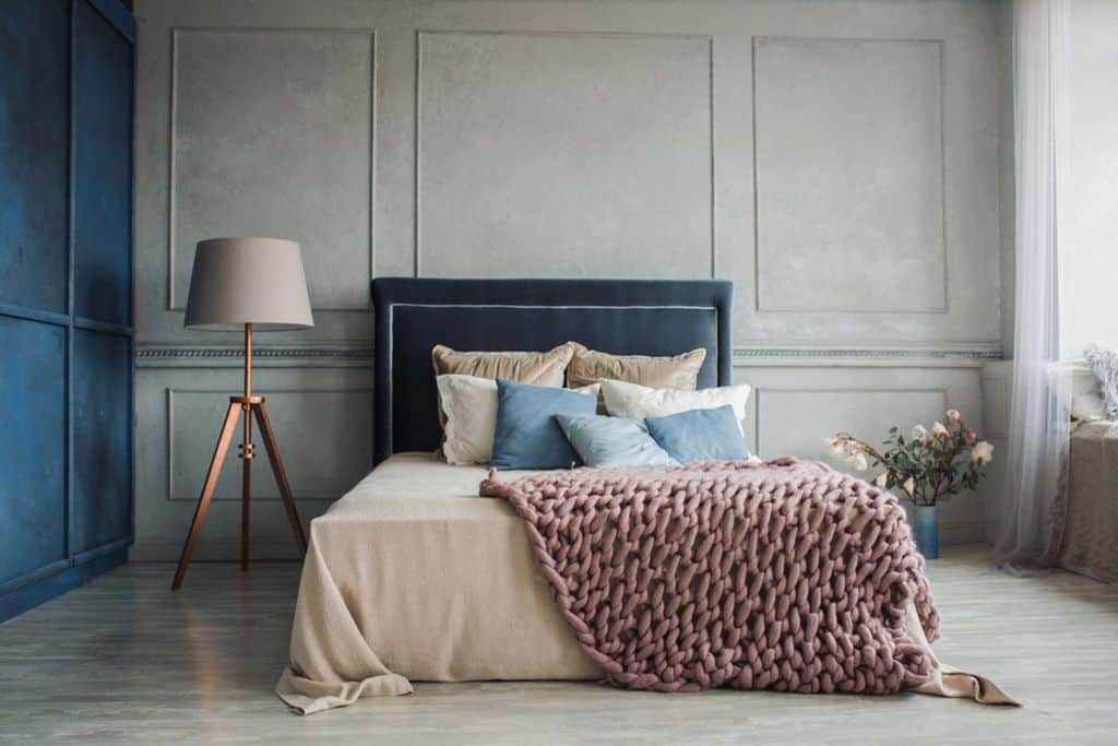 Minimalist bedroom with tufted headboard, knit blanket, bronze lamp, and floral accents.