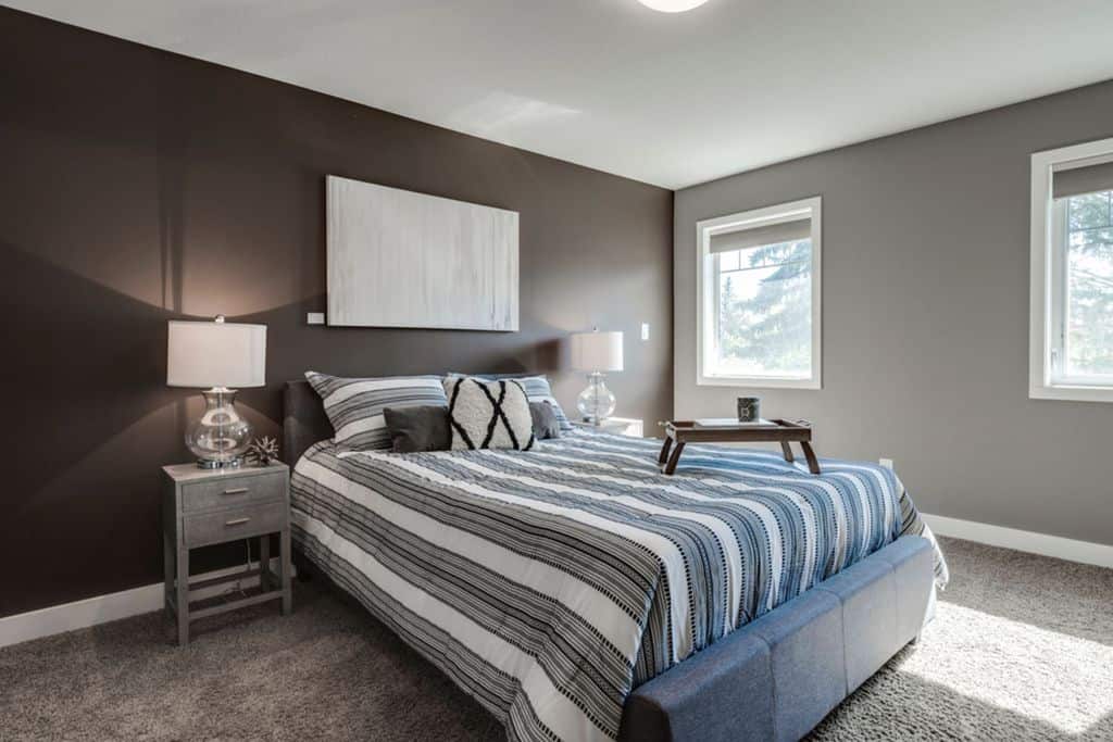 Modern bedroom with striped bedding, gray carpet, and natural light from two windows.