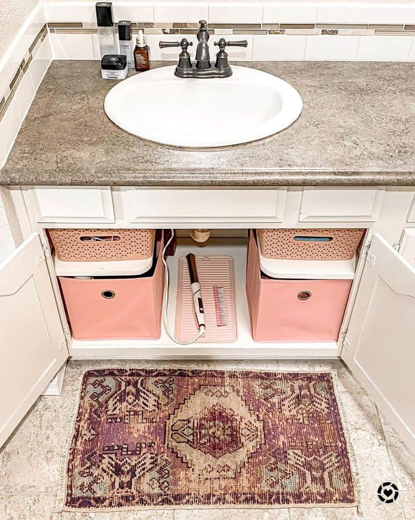 Bathroom sink cabinet with pink storage bins and countertop toiletries, with a patterned rug.