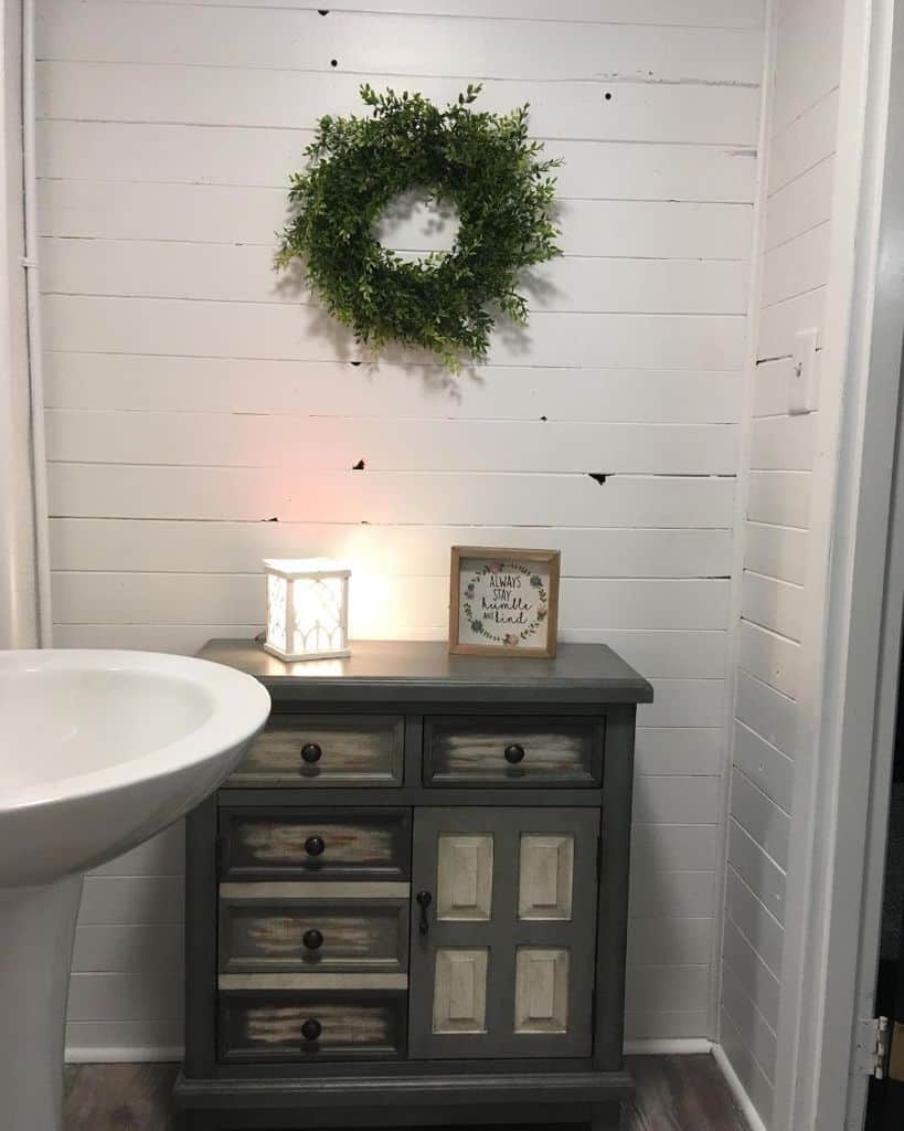 White bathroom with shiplap walls, rustic wood cabinet, and decorative wreath.