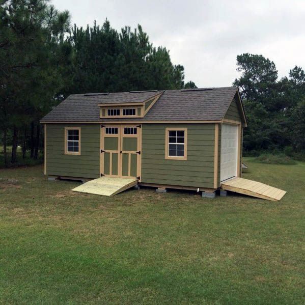 Green and beige backyard shed with farmhouse-style double doors and two ramps in an open field.