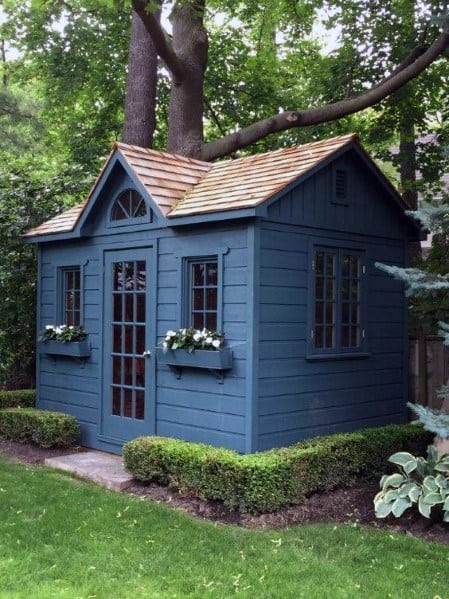 Blue wooden garden shed with French doors and flower boxes in a lush backyard.