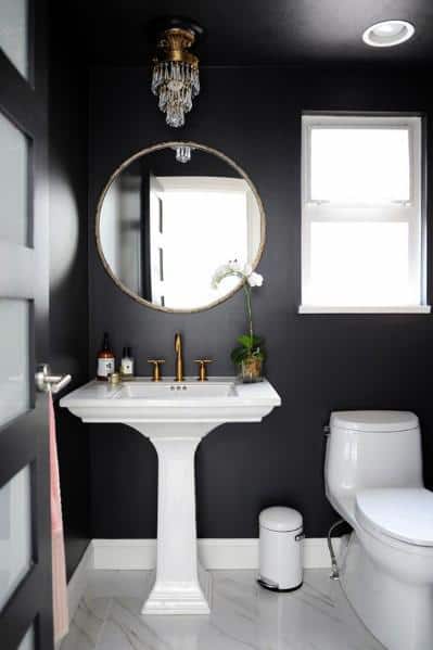 Black bathroom with pedestal sink, round gold mirror, chandelier, and natural light window.