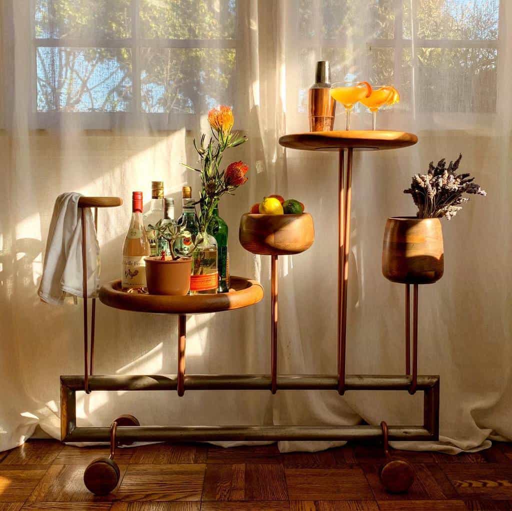 Unique wooden bar cart with various compartments holding drinks, fruit, and flowers in natural light.