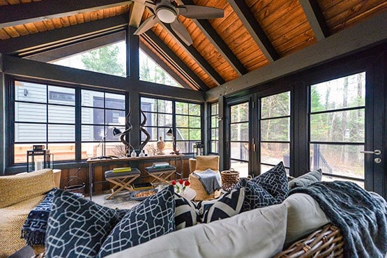 Rustic sunroom with vaulted wooden ceiling, black-framed windows, and cozy seating with patterned cushions.