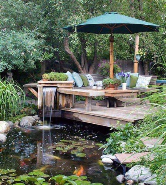 Wooden deck with seating area and umbrella, overlooking a koi pond with water plants and a small waterfall.