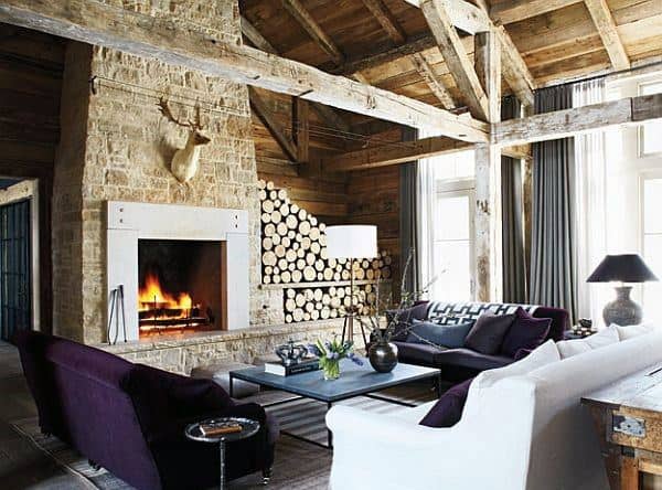 A unique rustic living room featuring a large stone fireplace, stacked wood, and a mix of luxurious seating with a warm and inviting ambiance