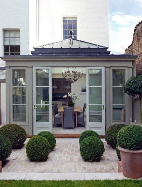 A charming sunroom with glass-paneled double doors, a pitched roof, elegant dining furniture, and a classic chandelier.