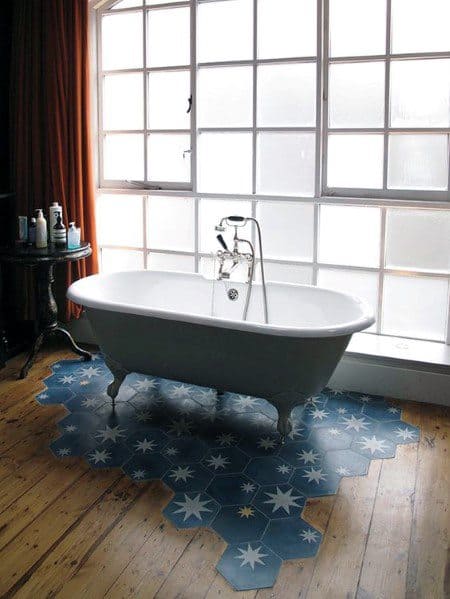 Freestanding bathtub on star-patterned blue tiles with wood floor transition and large frosted window.