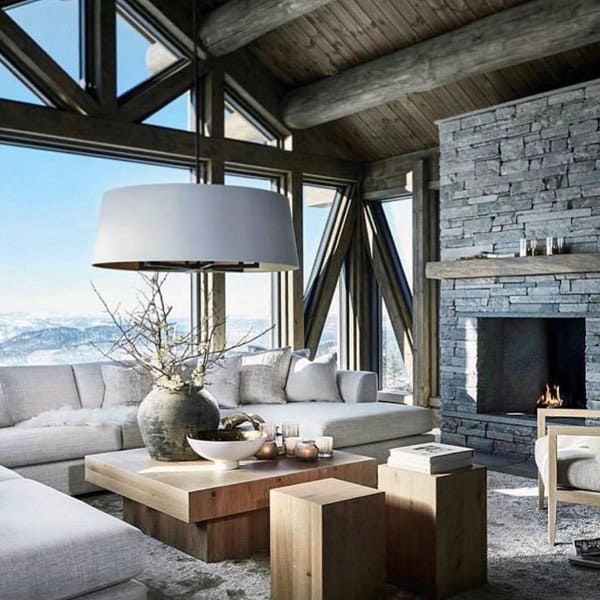 This rustic living room features a stunning stone fireplace, large windows offering breathtaking views, and a combination of cozy and modern furniture for a warm yet stylish ambiance. The use of wood and natural materials adds to the rustic charm while the open layout invites natural light to fill the space.