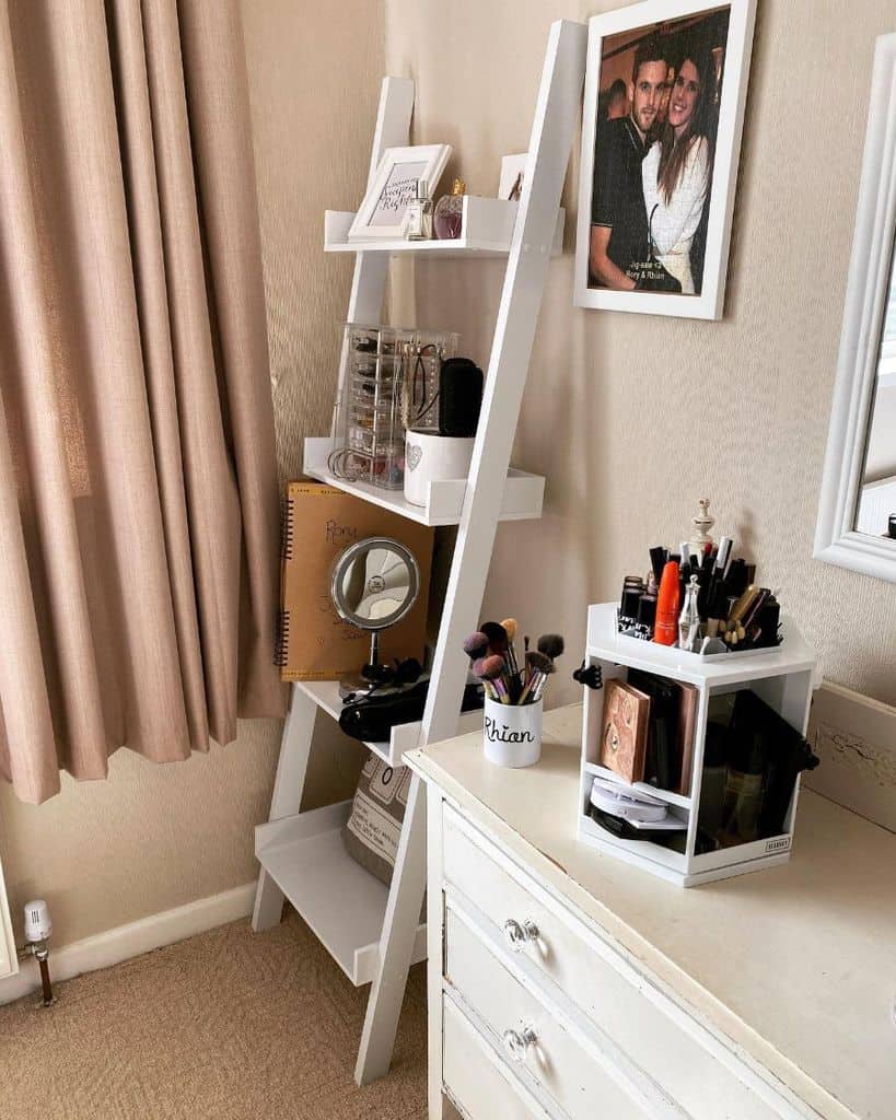 White ladder shelf with makeup, mirror, and decor next to white dresser in bedroom.