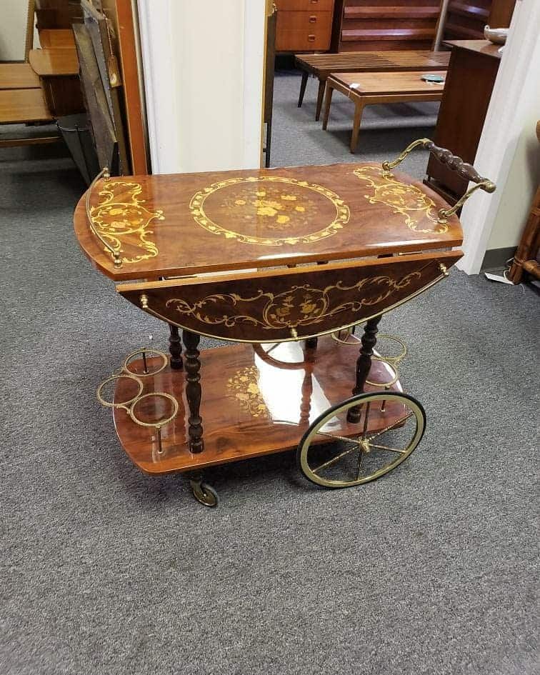 Vintage wooden bar cart with ornate gold detailing, drop-leaf sides, and large wheels.