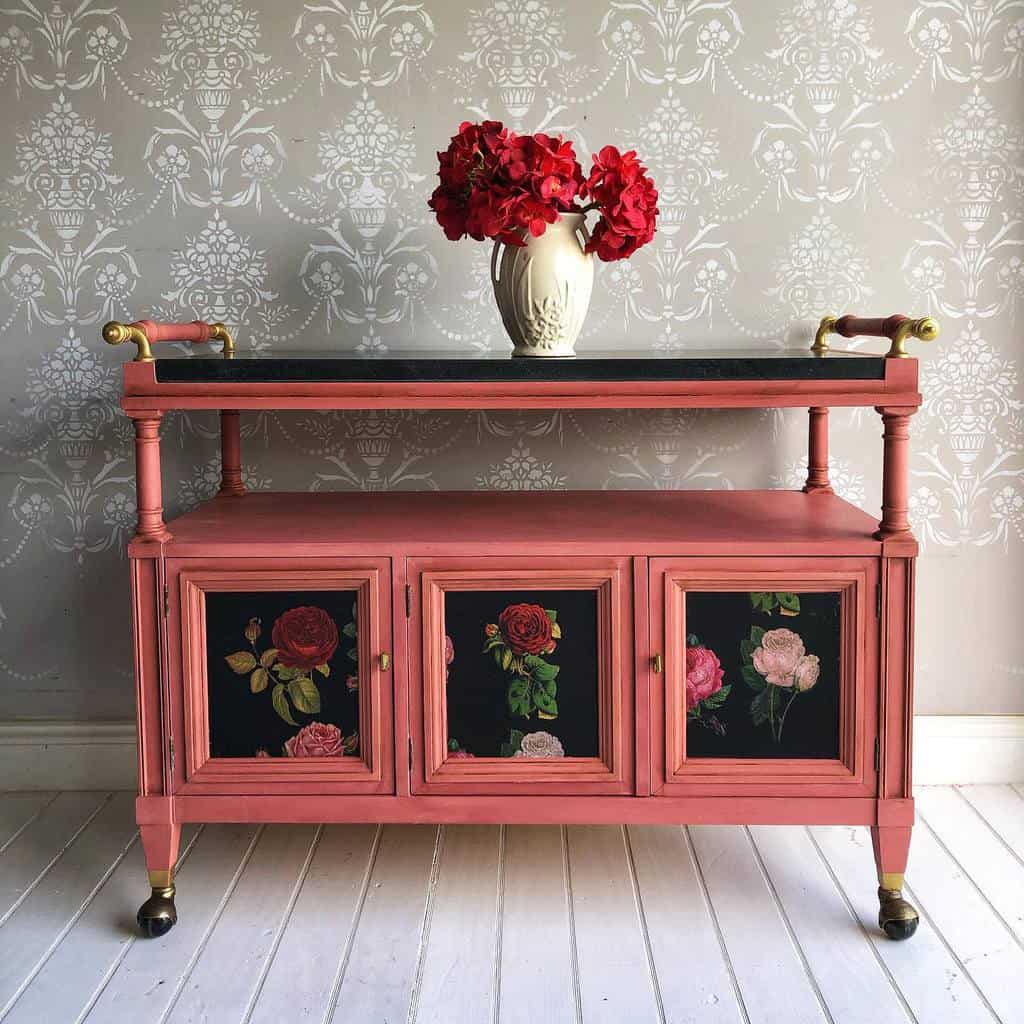 Pink bar cart with floral design on built-in drawers, gold handles, and a vase of red flowers.