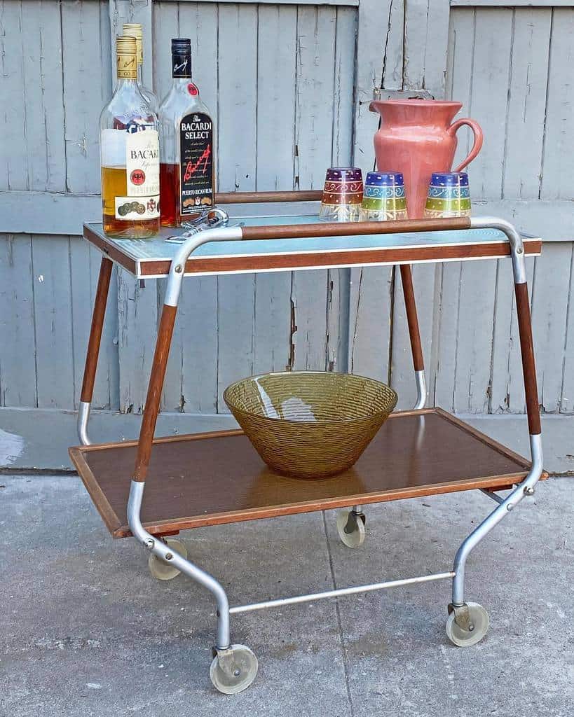 Vintage 70s bar cart with wooden shelves, liquor bottles, a pink pitcher, and colorful glasses.