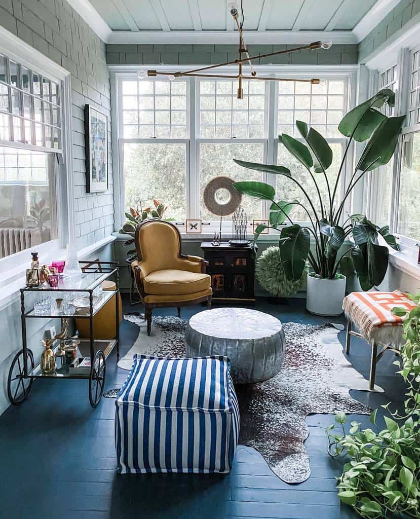 Vintage sunroom with a mustard armchair, lush greenery, a modern chandelier, and a striped ottoman for a cozy touch.