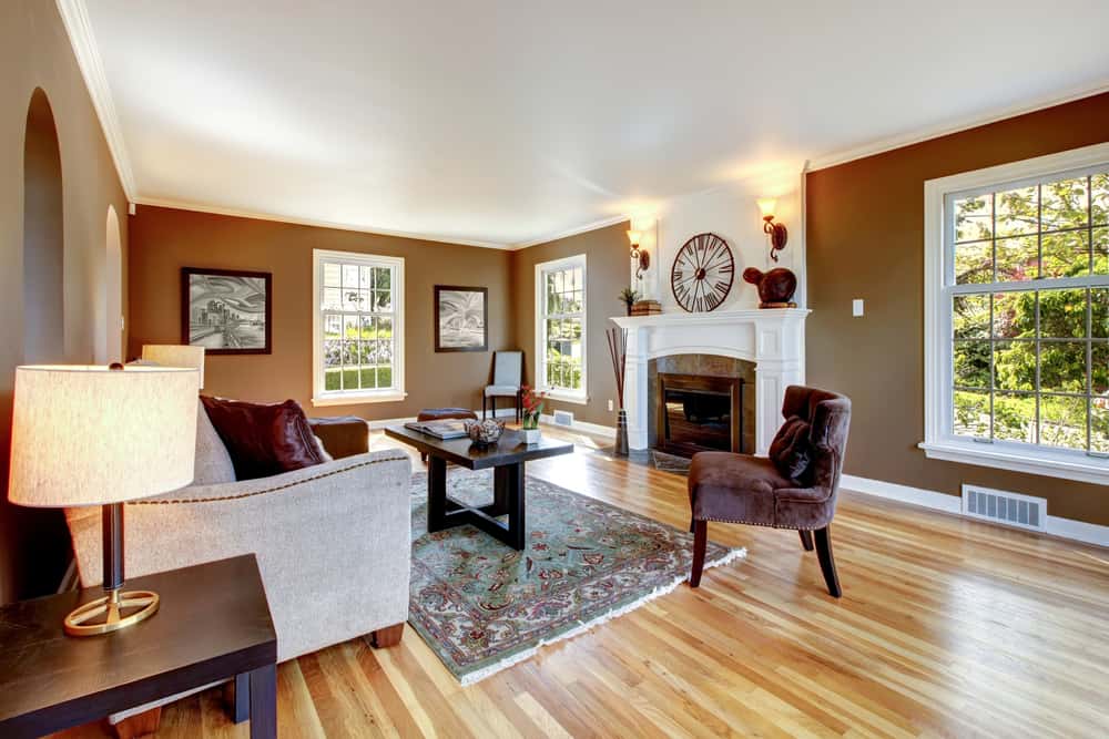 Cozy living room with brown walls, a sofa, armchairs, a fireplace, and large windows inviting natural light