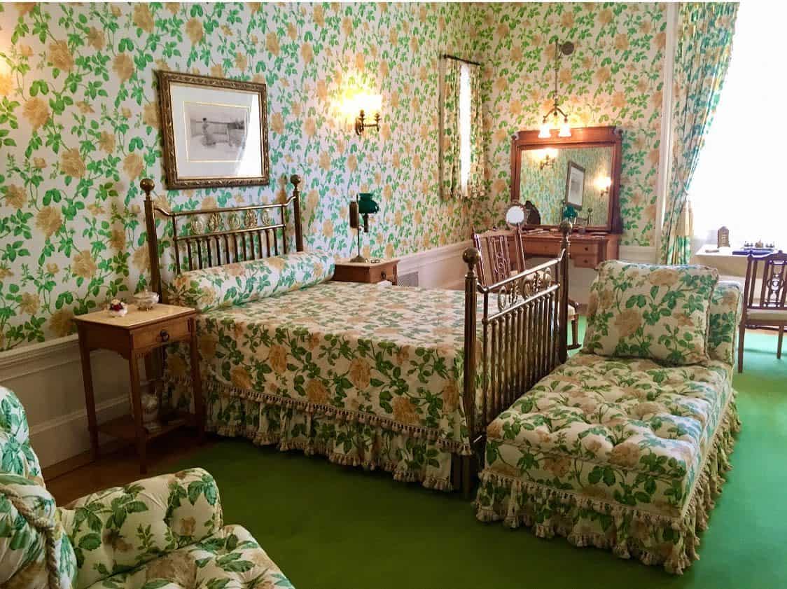 Vintage bedroom with floral wallpaper, matching bedspread and chaise, wooden furniture, and green carpet