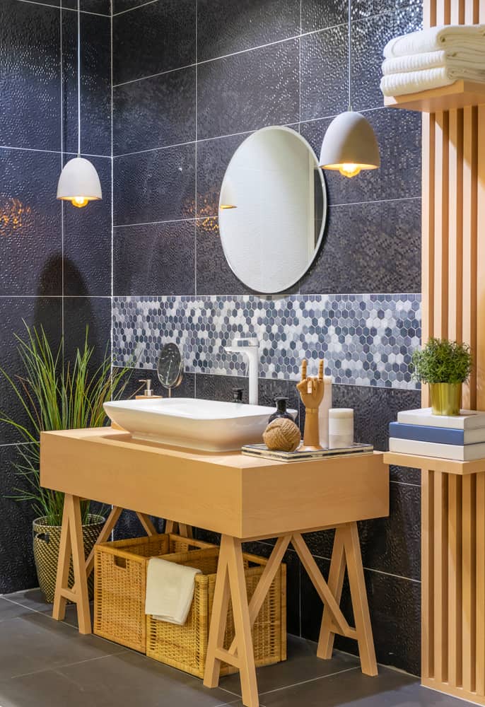 Modern bathroom with black tiled walls, wooden vanity, oval mirror, and hanging pendant lights.