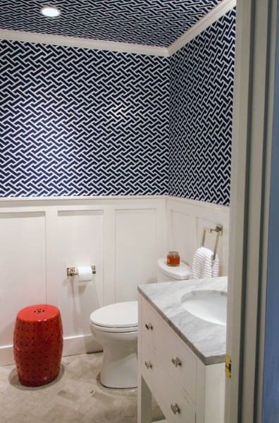 Small bathroom with blue geometric patterned wallpaper, white wainscoting, and red accent stool.