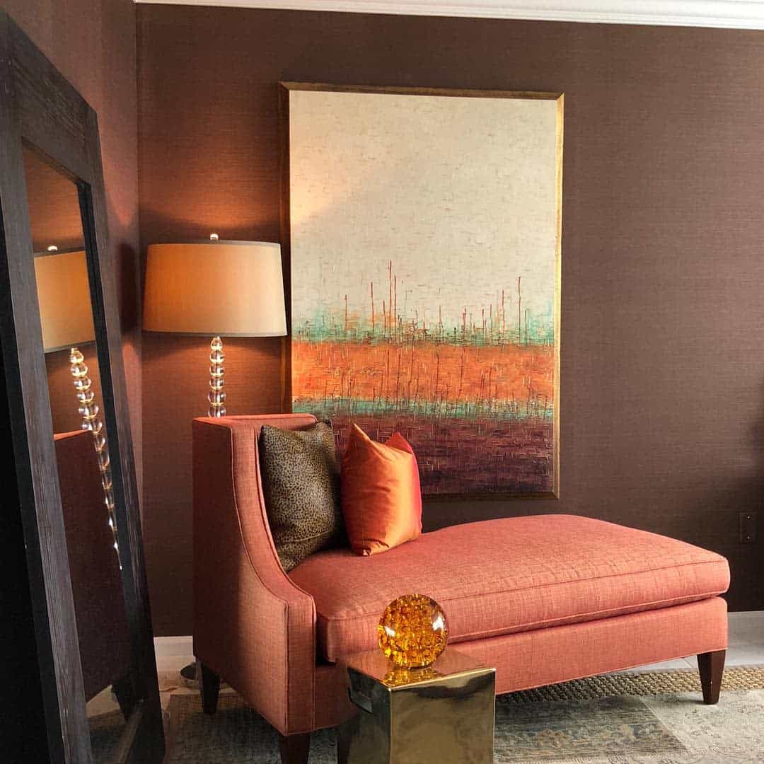 Room with an orange chaise lounge, brown abstract wall art, a lamp, and a mirror reflecting the scene