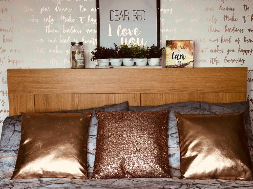 Bedroom with gold metallic script wallpaper, wooden headboard, and bronze cushions.