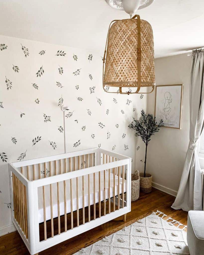 Minimalist baby room with white crib, leaf-patterned wallpaper, and woven pendant light.