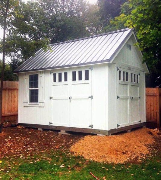 White barn-style shed with farmhouse doors, metal roof, and small windows in a fenced backyard.