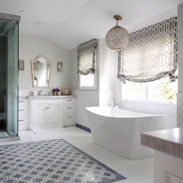 Freestanding white bathtub in an elegant bathroom with patterned window shades and chandelier.