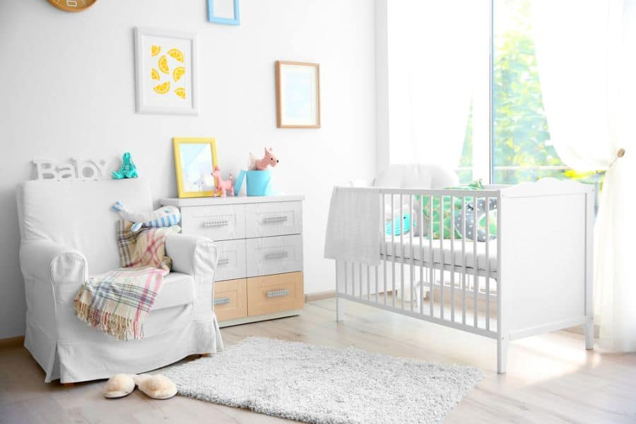 Bright baby room with a white armchair, crib, and pastel decor accents by a large window.