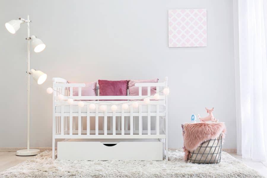 Baby room with white crib, pink pillows, string lights, and a cozy fur basket with a unicorn plush toy.