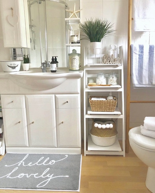 White bathroom with storage shelves, wicker basket, plants, and “hello lovely” rug in front.