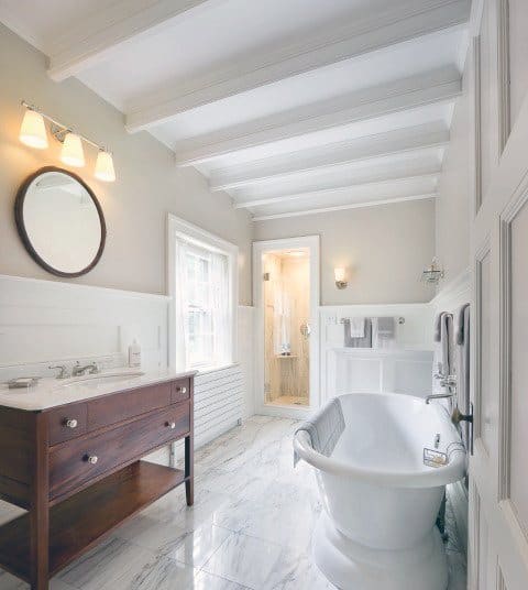 Bright bathroom with a clawfoot tub, wooden vanity, and a round mirror; marble floor and ceiling beams enhance the decor