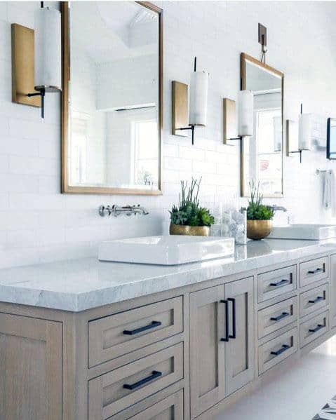 Light wood bathroom vanity with white countertop, brass mirrors, and white subway tile backsplash.