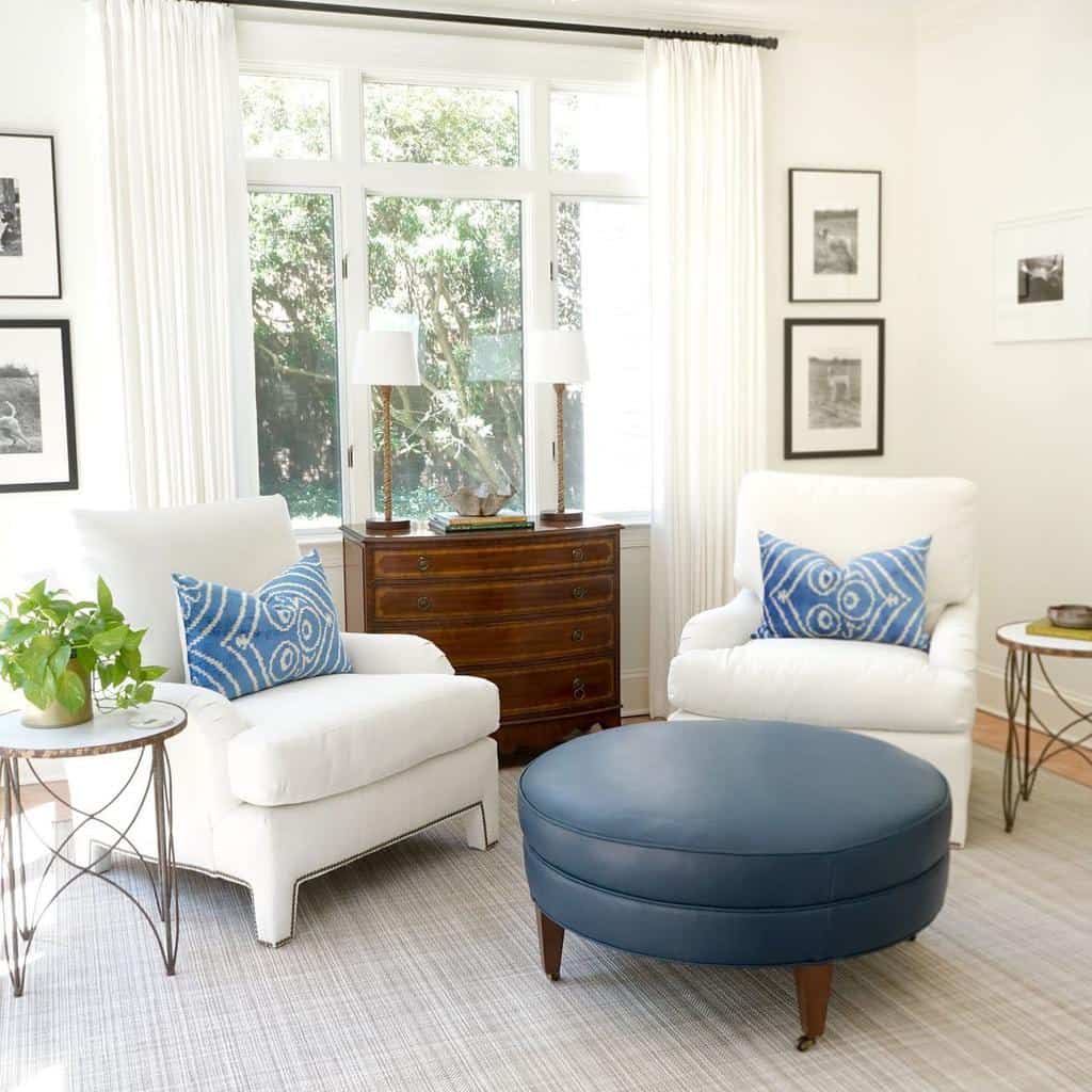 Sunroom with white armchairs, blue ottoman, wooden dresser, and decorative blue pillows.