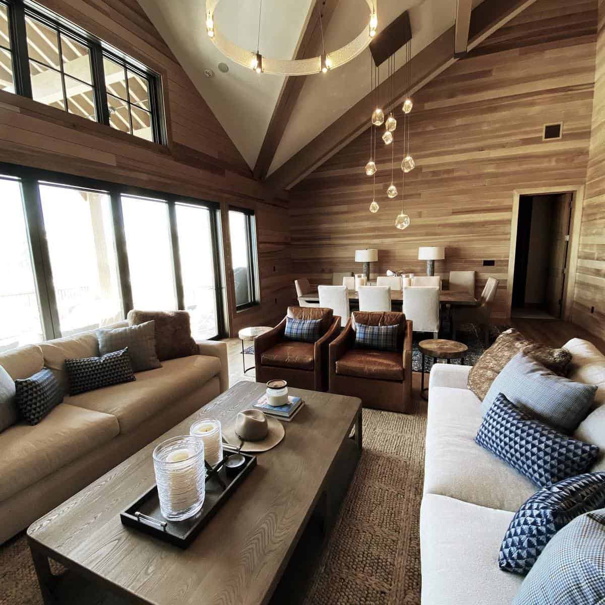 Cozy living room with large windows, brown wood paneling, sofas, armchairs, and a dining area with modern lighting