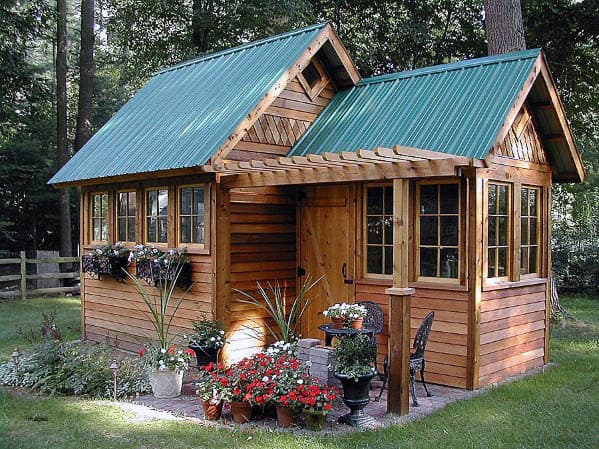 Rustic wood shed with green metal roof, multiple windows, and small garden patio.