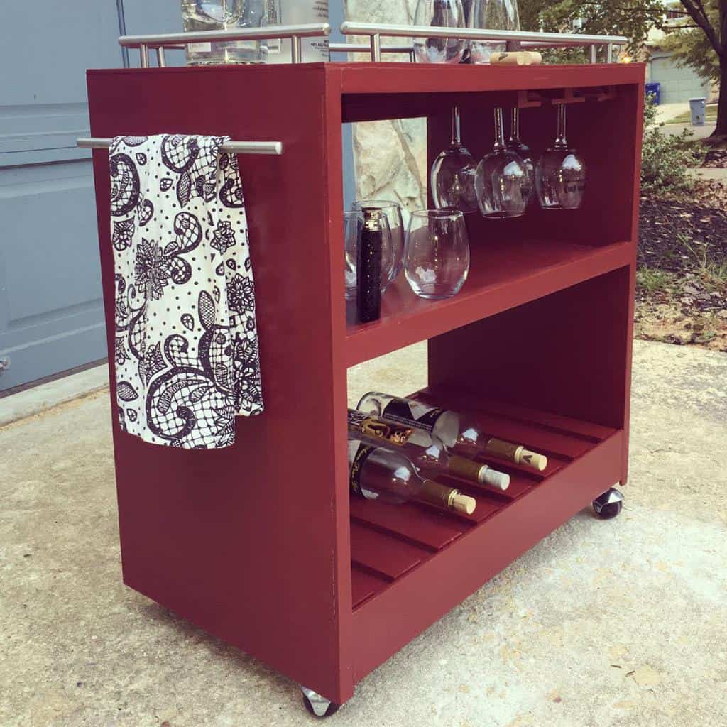 Simple red wooden bar cart with wine bottles, glassware, and a patterned towel hanging on the side.