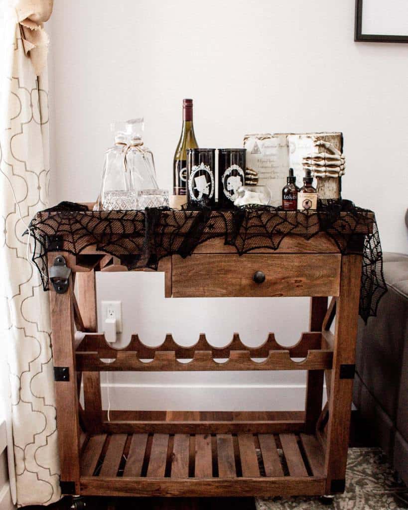 Wooden bar cart with wine rack, decorated with Halloween-themed items and a spiderweb tablecloth.