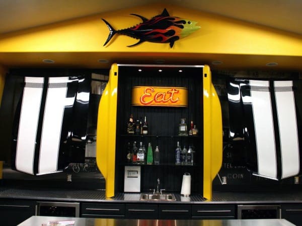 Modern garage bar with yellow car-themed decor, liquor shelves, and a neon "Eat" sign.