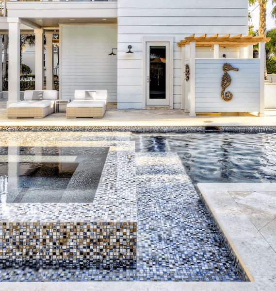 Modern pool with mosaic tile designs, seahorse decor, and white lounge chairs on a sunny patio.