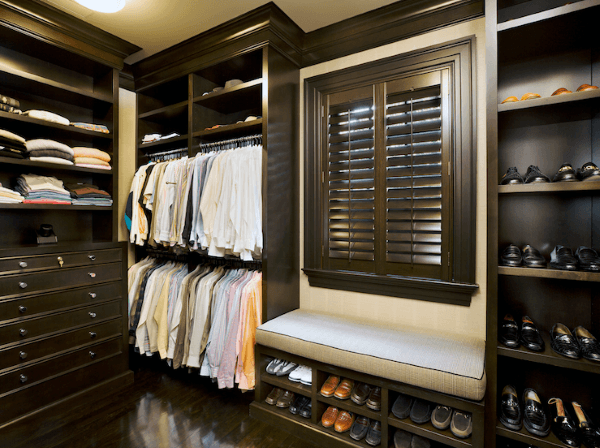 Walk-in closet with neatly arranged shirts, shoes, and a cushioned bench under shuttered window